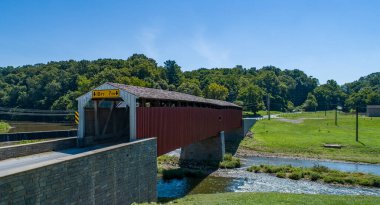 Tarihi örtülü bir köprü, berrak mavi gökyüzünün altında canlı yeşilliklerle çevrili, kırsal bir ortamda huzurlu bir nehri geçer. Huzurlu sahne ziyaretçileri keşfetmeye davet ediyor.