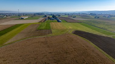 Geniş tarım arazileri ufuk boyunca uzanır ve öğle vakti açık mavi bir gökyüzü altında kahverengi, yeşil ve altın yama alanları sergiler. Traktörler ve çiftlik binaları manzarayı noktalıyor.