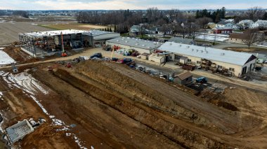 Kırsal bir alanda inşaat halindeki bir imalat tesisi, bölgede ağır makineler çalışıyor. Yakınlarda, tarım arazileri sanayi ve tarımın bir karışımını sergileyerek uzak mesafelere kadar uzanıyor..