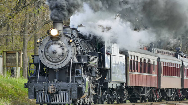 A vintage steam locomotive billows smoke as it rumbles through vibrant green fields under a bright blue sky, capturing the essence of a bygone era in rail travel.