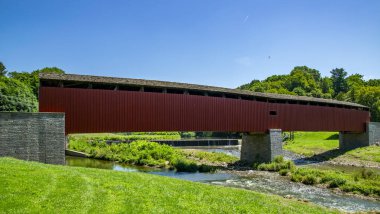 Çarpıcı kırmızı bir köprü yemyeşil bir çayırla çevrili yumuşak bir dereyi geçer. Güneş ışıl ışıl parlıyor, huzurlu kırsal bölgeye sıcak bir parıltı saçıyor..