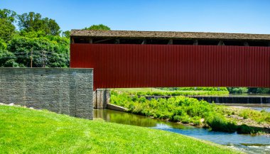 Canlı yeşil çimenler ve ağaçlarla çerçevelenmiş, dingin bir nehir boyunca uzanan kırmızı bir köprü. Açık mavi gökyüzü bu sakin manzaranın resimsel görüntüsünü güçlendiriyor..
