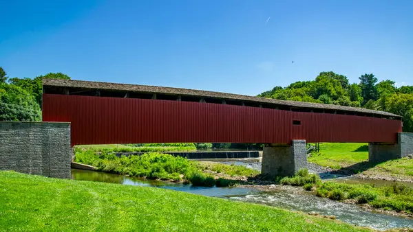 Çarpıcı kırmızı bir köprü yemyeşil bir çayırla çevrili yumuşak bir dereyi geçer. Güneş ışıl ışıl parlıyor, huzurlu kırsal bölgeye sıcak bir parıltı saçıyor..