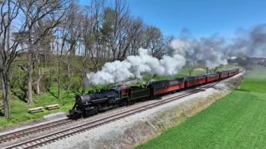 Ronks, Pennsylvania, 9 Nisan 2025 - Buharlı bir lokomotif yemyeşil tarlalar ve ağaçlarla çevrili raylar boyunca klasik bir yolcu treni çekiyor. Güneş ışıl ışıl parlıyor, resim gibi bir manzara yaratıyor.