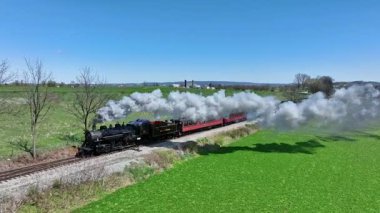 Ronks, Pennsylvania, 9-2025 tarihsel bir buhar lokomotifi raylar boyunca hareket ediyor ve buhar bulutları salıyor. Bereketli yeşil tarlalar ve açık mavi bir gökyüzü resimsel bir manzara yaratır