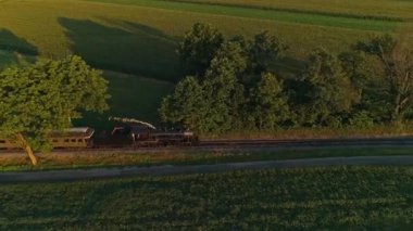 Klasik bir buhar lokomotifi, gün batımında yemyeşil tarlalar ve ağaçlarla çevrili dolambaçlı bir yol boyunca vagonları çeker. Kırsal kesimin sakinliği tam ekrana yansıyor..