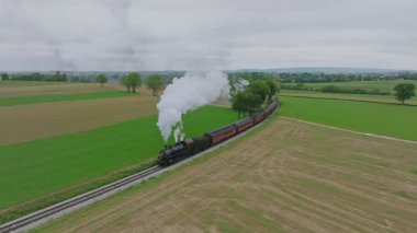 Muhteşem bir buhar treni, yemyeşil çimenliklerle çevrili görkemli tarlalar boyunca tıkınıyor. Nazik duman, sakin manzaraya doğru ilerlerken arkasında iz bırakır..