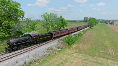 Ronks, Pennsylvania, 5 Mayıs 2023 - Buharlı bir tren açık mavi gökyüzünün altında yemyeşil alanlarda süzülüyor. Çekici lokomotif yolcuları taşırken doğanın güzelliğini gösteriyor..