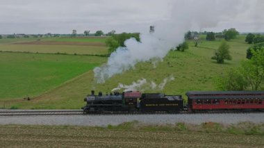 Ronks, Pennsylvania, 5 Mayıs 2023 - Yeşillik yeşil tarlalar ve ağaçlarla çevrili demiryolu rayları boyunca ilerleyen klasik bir buhar lokomotifi. Motordan dumanlar yükseliyor ve geçmişe bir göz atıyor.