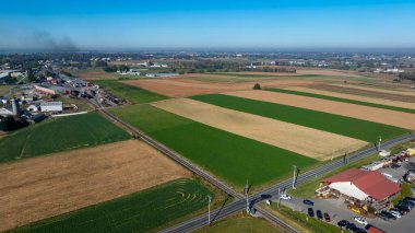 Değişik yeşil ve kahverengi tarlaları olan canlı tarım arazilerine geniş bir bakış açısı. Bölgeden geçen sakin bir kırsal yol, küçük bir kasaba ve açık gökyüzünün altındaki binalarla sınırlıdır..