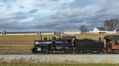 Klasik bir buhar treni bulutlu bir gökyüzünün altında geniş tarlalar boyunca akar ve kırsal alanda nostaljik bir yolculuk sergiler. Tren eski moda bir cazibe yayar..