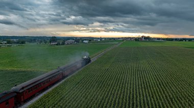 Kaliteli bir buhar treni, yemyeşil mısır tarlaları tarafından kenarlı raylar boyunca hareket eder. Güneş batarken tepemizde kara bulutlar beliriyor. Huzurlu kırlara altın bir parıltı saçıyor..