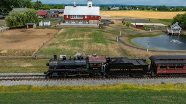 Ronks, Pennsylvania, 11 Temmuz 2024 - Klasik bir buhar treni yemyeşil alanlar boyunca bir yolcu vagonunu çeker. Yakınlarda büyüleyici kırmızı bir ahır var. Yuvarlanan tepeleri ve görülebilen bir göleti var.