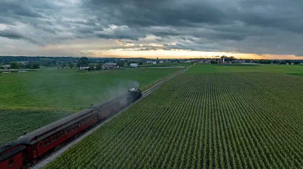 Kaliteli bir buhar treni, yemyeşil mısır tarlaları tarafından kenarlı raylar boyunca hareket eder. Güneş batarken tepemizde kara bulutlar beliriyor. Huzurlu kırlara altın bir parıltı saçıyor..