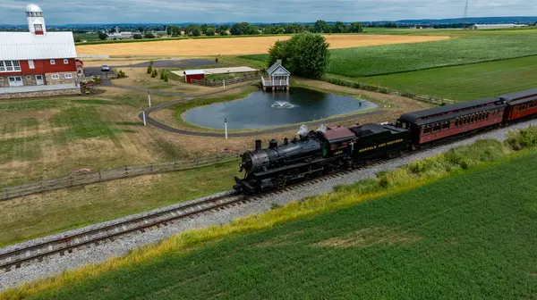 Ronks, Pennsylvania, 11 Temmuz 2024 raylar boyunca yemyeşil alanlardan ve sakin bir göletten geçen klasik bir buhar lokomotifi. Parlak kırmızı ahır yakınlarda duruyor, cennet bahçesini tamamlıyor.