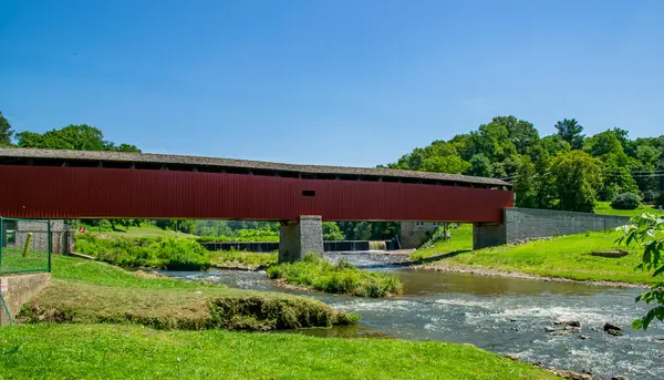 Kırmızı kaplı bir köprü, parlak ve güneşli bir günde canlı yeşil ağaçlarla çevrili yumuşak bir derenin üzerinden geçer. Doğa bu barışçıl kırsal alanda gelişiyor, keşif yapmaya davet ediyor..