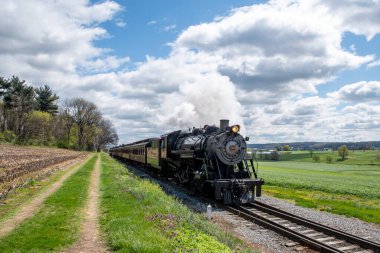 Ronks, Pennsylvania, 16 Nisan 2025 - Buharlı bir lokomotif yeşil alanlara bitişik demiryolu rayları boyunca süzülürken beyaz duman püskürtür. Manzara parçalı bulutlu gökyüzü altında canlı bahar yapraklarına sahiptir.,