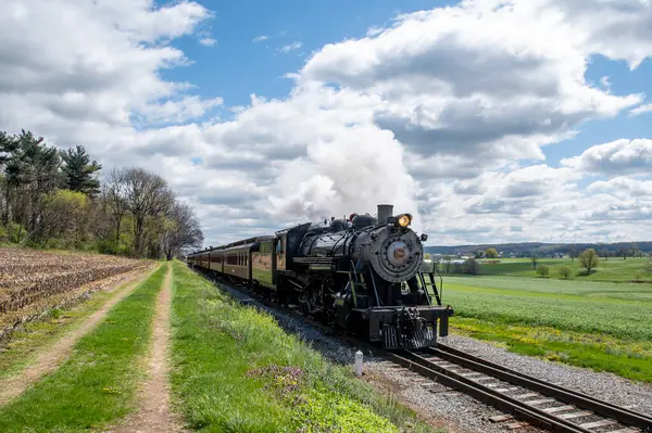 Ronks, Pennsylvania, 16 Nisan 2025 - Buharlı bir lokomotif yeşil alanlara bitişik demiryolu rayları boyunca süzülürken beyaz duman püskürtür. Manzara parçalı bulutlu gökyüzü altında canlı bahar yapraklarına sahiptir.,