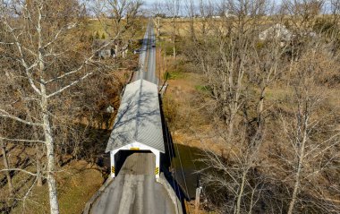 Çıplak ağaçlar ve sonbahar yapraklarıyla çevrili huzurlu bir derenin üzerinde uzanan bir köprü. Açık gökyüzü ve yumuşak ışık huzurlu atmosferi güçlendirir..