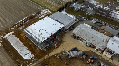 İşçiler büyük bir ticari binanın çatısında meşgulken, ağır makineler ve malzemeler sahanın etrafına saçıldı. Kırsal bir alanda kar zeminin bazı kısımlarını kaplar..