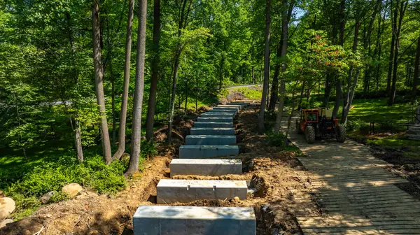 Bir inşaat alanı, verimli bir ormanlık alanda yürüme yolu oluşturmak için beton döşeyen işçileri gösteriyor. Ağır makineler mevcuttur ve çevreyi çevreleyen ağaçlar doğal manzarayı güçlendirir..