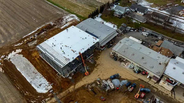 İşçiler büyük bir ticari binanın çatısında meşgulken, ağır makineler ve malzemeler sahanın etrafına saçıldı. Kırsal bir alanda kar zeminin bazı kısımlarını kaplar..