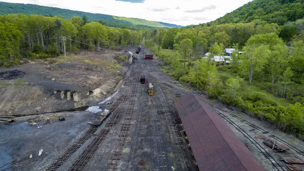 Terkedilmiş demiryolu alanının geniş bir manzarası. Aşırı gelişmiş rayları ve terk edilmiş tren araçlarının kalıntıları var. Verdant ağaçları bulutlu bir gökyüzünün altındaki yuvarlanan tepelerin zeminine karşı bölgeyi sınırlandırır.