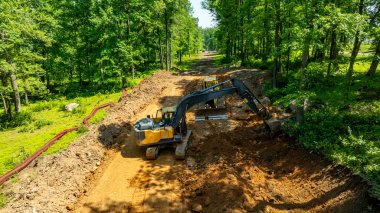 Kazıcılar yoğun bir ormanlık alandaki araziyi temizlemeye çalışıyorlar. Operatörler, bölgeyi açık mavi gökyüzü altında gelecekteki gelişime hazırlamak için ağır ekipmanlar kullanıyorlar..