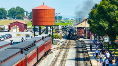 Strasburg, Pennsylvania, 26 Ağustos 2024 - Hevesli ziyaretçiler, klasik bir buharlı trenin durduğu pitoresk bir tren istasyonunda toplandılar. Bereketli tarlalar ve su kulesi açık bir yaz gününde büyüleyici bir zemin oluşturur..