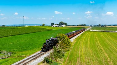 Buharlı bir lokomotif geniş yeşil alanlar ve verimli tarım arazileri boyunca uzun bir tren çeker. Parlak gökyüzü ve uzak çiftlikler kırsal bir manzara yaratıyor..