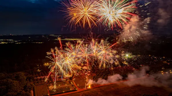 Havai fişekler, ağaçlar ve kalabalıkla çevrili kırsal bir ortamda genişleyen bir açık hava olayının üzerindeki karanlık gökyüzünü aydınlatır. Parlak renkler patlıyor ve gece boyunca neşeli bir atmosfer yaratıyor..