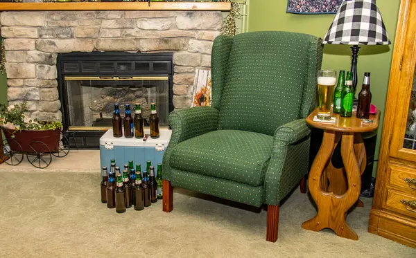 Sıcak bir oturma odasında, taş bir şöminenin yanında yeni dökülmüş birayla döşenmiş yeşil bir sandalye yer alır. Dolu bira şişeleri etrafa saçılmış durumda.