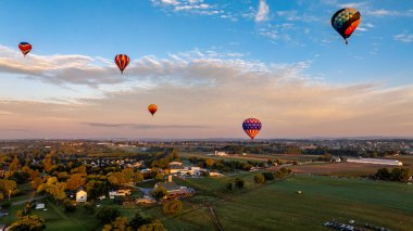 Sıcak hava balonları zarif bir şekilde gökyüzüne yükselir, günbatımında sakin bir kırsal ortamın arka planına canlı renkler saçar, uçuşun ve doğanın güzelliğini vurgular..