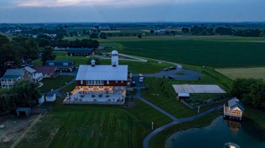 Akşamları sıcacık yanan kırsal bir ahır geniş yeşil tarlaların arasında yer alır. Yakınlarda, sakin bir gölet güneş batarken parlayan ışıkları yansıtıyor ve sakin bir atmosfer yaratıyor..
