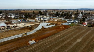 Ağır makineler toprağı derecelendirme ve kırsal bir alanda yeni bir kalkınma projesine hazırlanma üzerinde çalışıyor. Manzara, evler, açık alanlar ve arka planda görünen uzak bir kasabayı içerir..