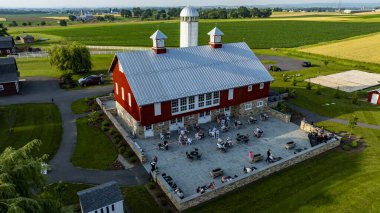 Ronks, Pennsylvania, 12 Haziran 2025 - Yeşil tarlalara karşı büyüleyici bir kırmızı ahırda canlı bir toplantı gerçekleşti. Ziyaretçiler terasta dinleniyor, sıcak bir kırsal atmosferin tadını çıkarıyorlar.