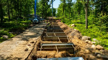 İşçiler ağaçlık bir bölgede kamu siperleri inşa etmekle meşguller. Ağır makineler yakınlarda park halinde ve bölge yemyeşil ağaçlar ve kayalarla çevrili..