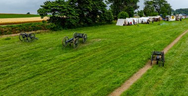 Topun kopyaları çimenli bir alana yerleştirilirken, yakınlardaki tuvaller canlı bir atmosfer yaratıyor. Katılımcılar tarihi olayları anımsatan etkinliklere katılıyorlar.