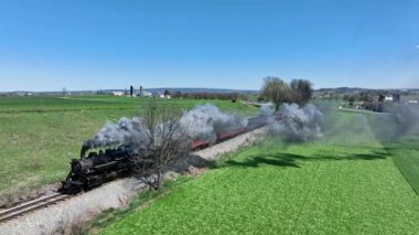 Ronks, Pennsylvania, 9-2025 - Canlı yeşil alanlar ve ağaçlarla çevrili manzaralı raylar boyunca klasik bir buhar lokomotifi. Gökyüzü açık, güneşli güzel bir gün sergileniyor..