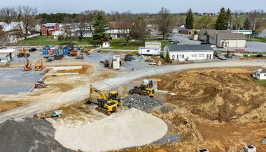 Ağır makineler büyük bir inşaat sahasında çalışır, toprak hareket ettirilir ve malzemeler düzenlenir. Yakınlardaki binalar ve yeşil alanlar, devam eden kalkınmaya zemin hazırlıyor.