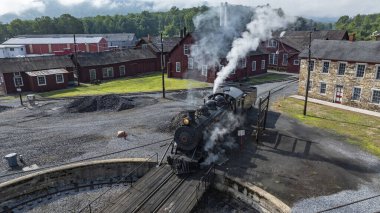 Kırmızı ve taştan binalarla çevrili bir demiryolu müzesinde tarihi bir buhar treni ahşap bir platforma dönüyor. Lokomotiften gelen buhar dalgaları atmosferik bir manzara yaratıyor..