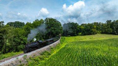Tarihi bir buhar treni dolambaçlı bir yol boyunca tıkırdıyor. Etrafı yemyeşil tarlalar ve parlak mavi gökyüzünün altındaki ağaçlarla çevrili..