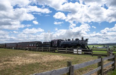 Buhar lokomotifi bulutlarla dolu açık mavi bir gökyüzünün altında yeşil bir arazi boyunca klasik tren vagonları çeker. Yakınlardaki ahşap çitler rayları çiziyor, kırsal çekiciliği arttırıyor..