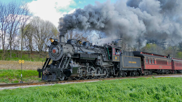 Ronks, Pennsylvania, April 13, 2025 - Historic steam locomotive travels along scenic railway, releasing clouds of smoke into the air. The train is surrounded by greenery, and vibrant spring landscape.