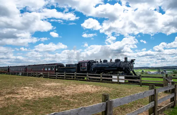 Buhar lokomotifi bulutlarla dolu açık mavi bir gökyüzünün altında yeşil bir arazi boyunca klasik tren vagonları çeker. Yakınlardaki ahşap çitler rayları çiziyor, kırsal çekiciliği arttırıyor..