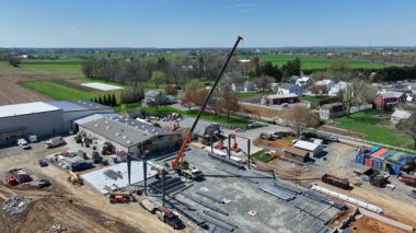 Kırsal kesimde yeşil tarlaları ve dağınık evleri olan çelik kirişleri kaldıran bir vince sahip bir inşaat alanının insansız hava aracı görüntüleri büyüleyici görüntüler, canlı renkler, pürüzsüz hareketler.