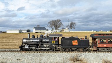 Ronks, Pennsylvania, 29 Aralık 2024 - Bir buhar lokomotifi geniş tarlalar ve uzak bir çiftlik evi ile çevrili raylar boyunca ilerliyor. Tren yolculuğunu yaparken tepemizde gri bulutlar sallanıyor.