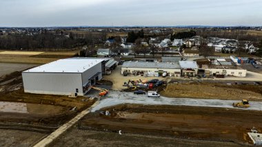 Kırsal bir yerde büyük bir depo inşa ediliyor. Ağır makineler ve araçlar mevcutken, yakınlardaki evler bir banliyö arka planı ekliyor. Çalışma alanı hareketlilikle meşgul görünüyor.