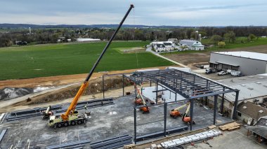 Vinçler ve inşaat ekipmanları kırsal bir bölgedeki yeni bir binanın çerçevesi üzerinde aktif olarak çalışıyor. Manzara, devam eden gelişmeleri gösteren alanlar ve yakınlardaki yapıları içerir..