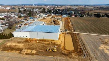 Konut evleri ve tarım arazileriyle çevrili kırsal bir alanda büyük bir sanayi yapısı inşa ediliyor. Bölgede etkin gelişim gösteren inşaat araçları görülüyor.